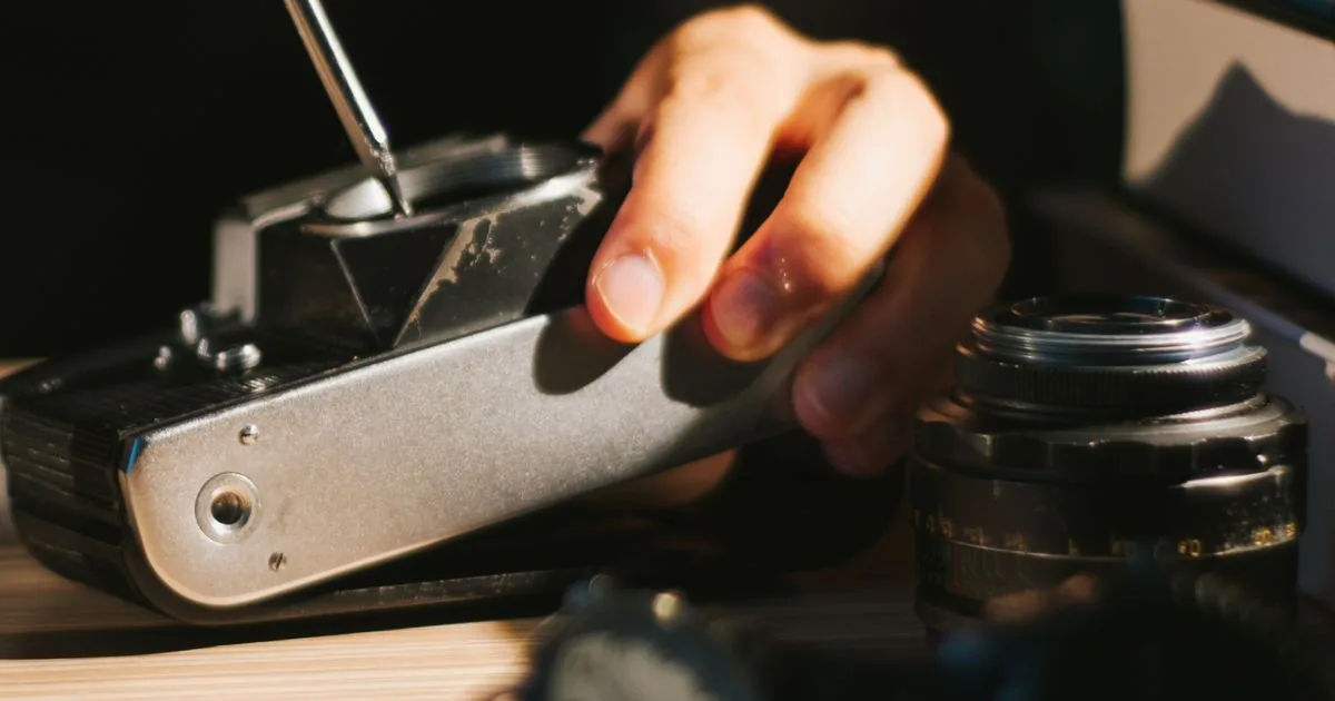 A technician's hand uses a screwdriver on the open top of a vintage film camera on a wooden workbench, with a lens nearby