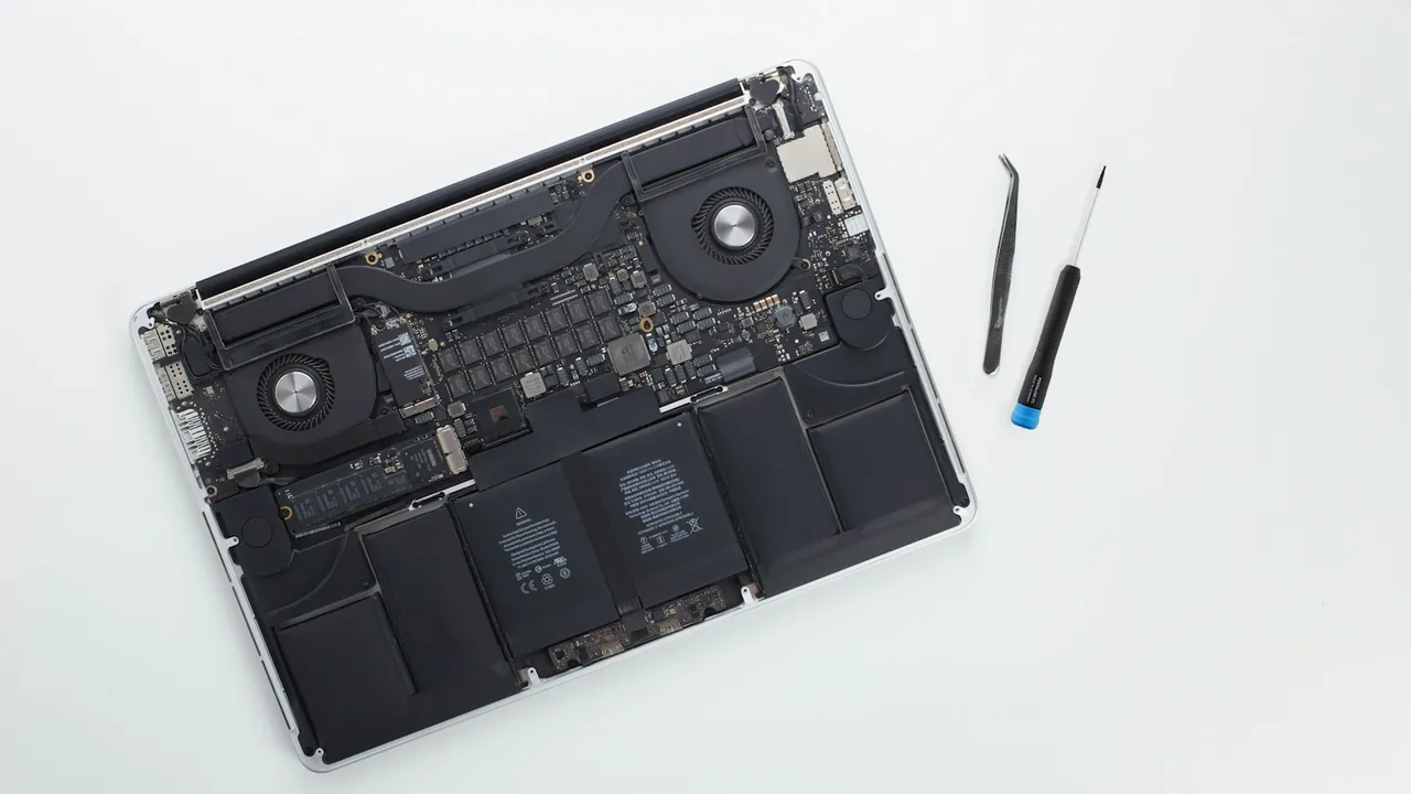 Laptop with its bottom cover removed and internal components exposed, lying on a white surface beside a small screwdriver and tweezers.
