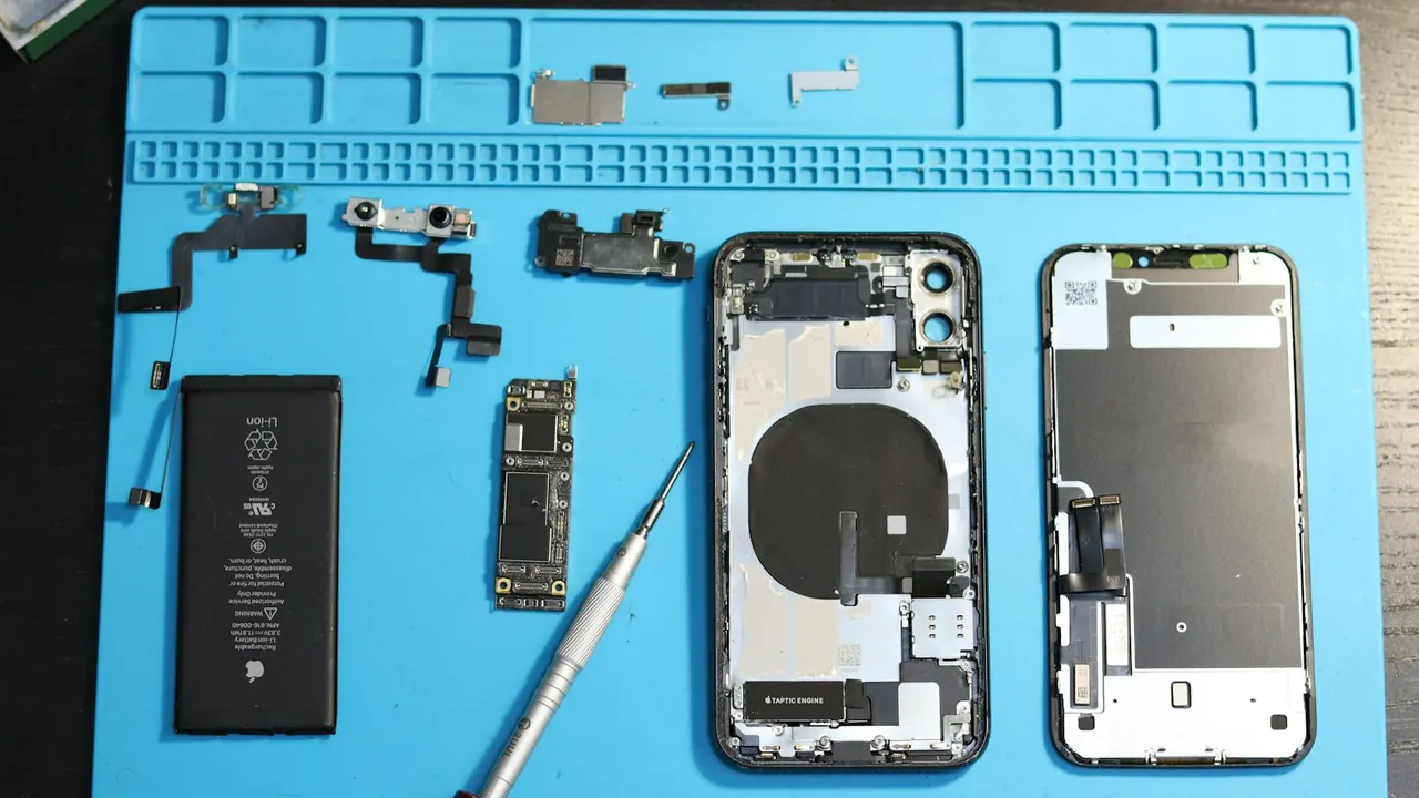 Disassembled phone parts, including the battery, logic board, camera and screen, laid out in order on a blue repair mat beside a screwdriver.