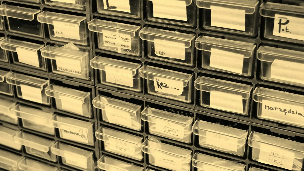 Rows of small clear plastic parts drawers with handwritten paper labels fill a metal storage cabinet.