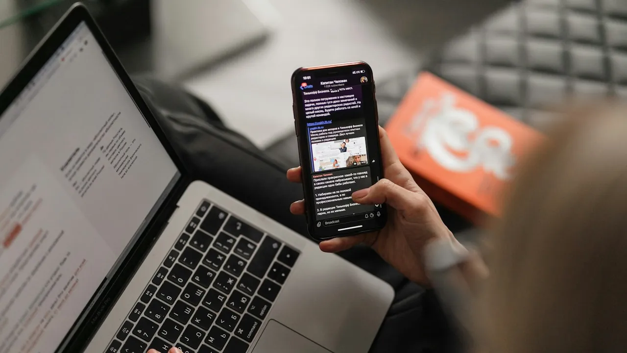 A hand holds a smartphone showing a text web page, next to an open laptop on a lap.