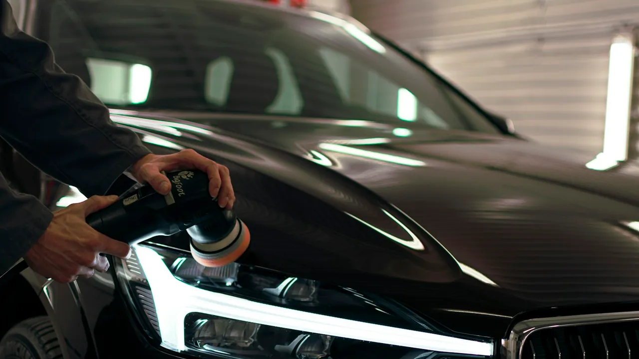 A detailer holds a orange-pad machine polisher against the hood of a glossy black car inside a garage.